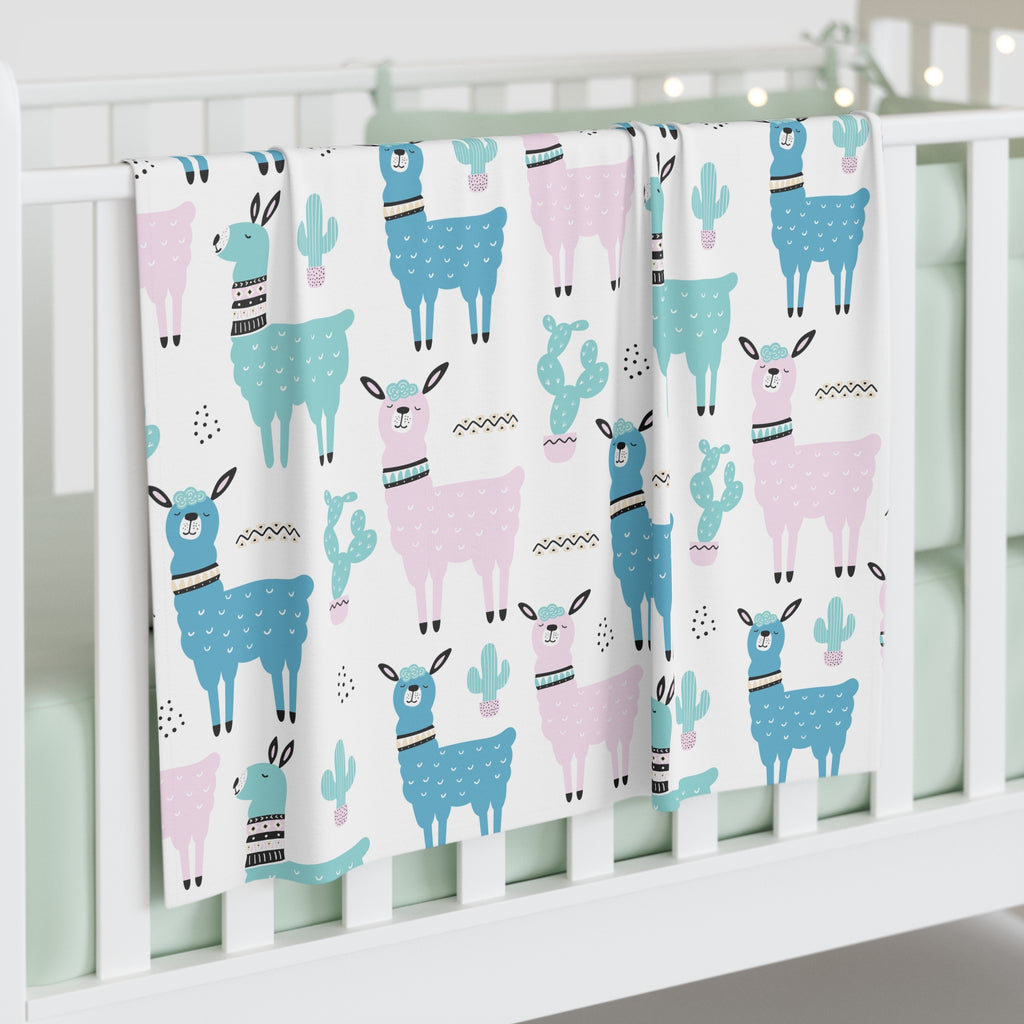 A white baby crib blanket featuring a playful pattern of blue and pink llamas and cacti, draped over the side of a white wooden crib.