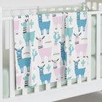 A white baby crib blanket featuring a playful pattern of blue and pink llamas and cacti, draped over the side of a white wooden crib.