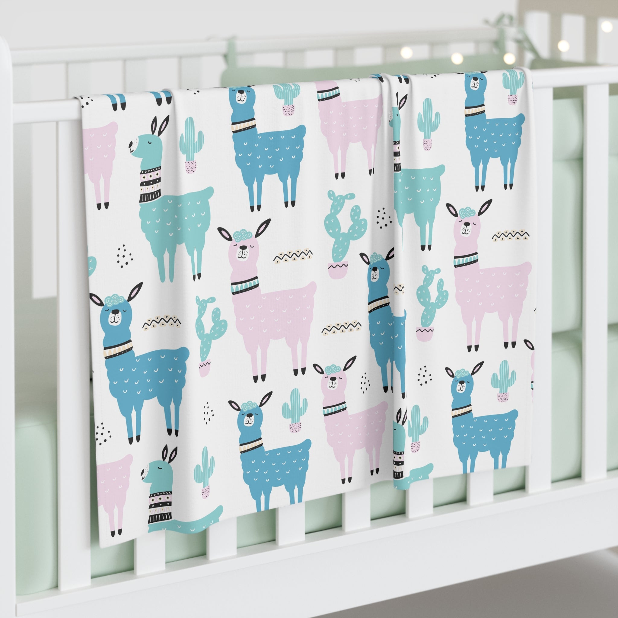 A white baby crib blanket featuring a playful pattern of blue and pink llamas and cacti, draped over the side of a white wooden crib.